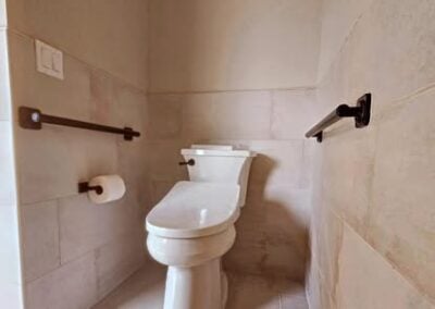 Modern bathroom with a white toilet, brown grab bars, and light-colored tiled walls, showcasing a clean and functional design for remodeling inspiration.